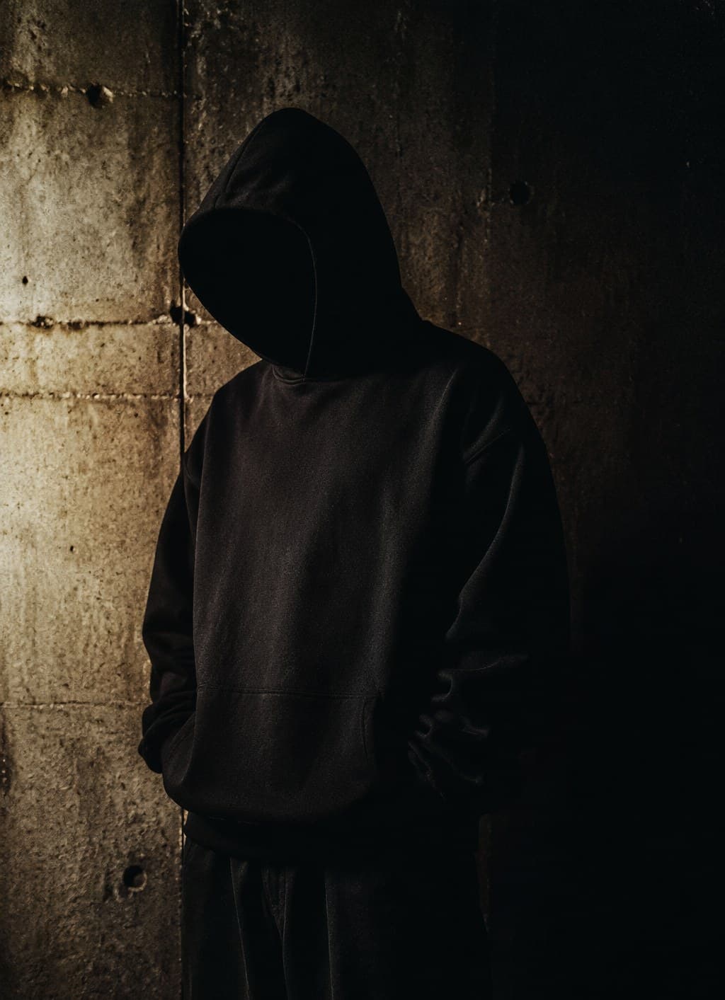 Hooded figure against a dark concrete wall, showing the boxy structured fit of the black hoodie